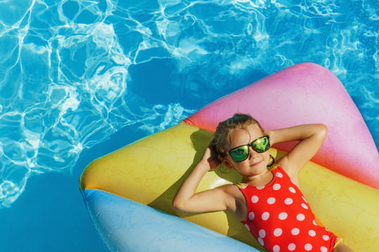 Kind mit Sonnenbrille liegt auf Luftmatratze im Pool und verschränkt die Arme hinter dem Kopf